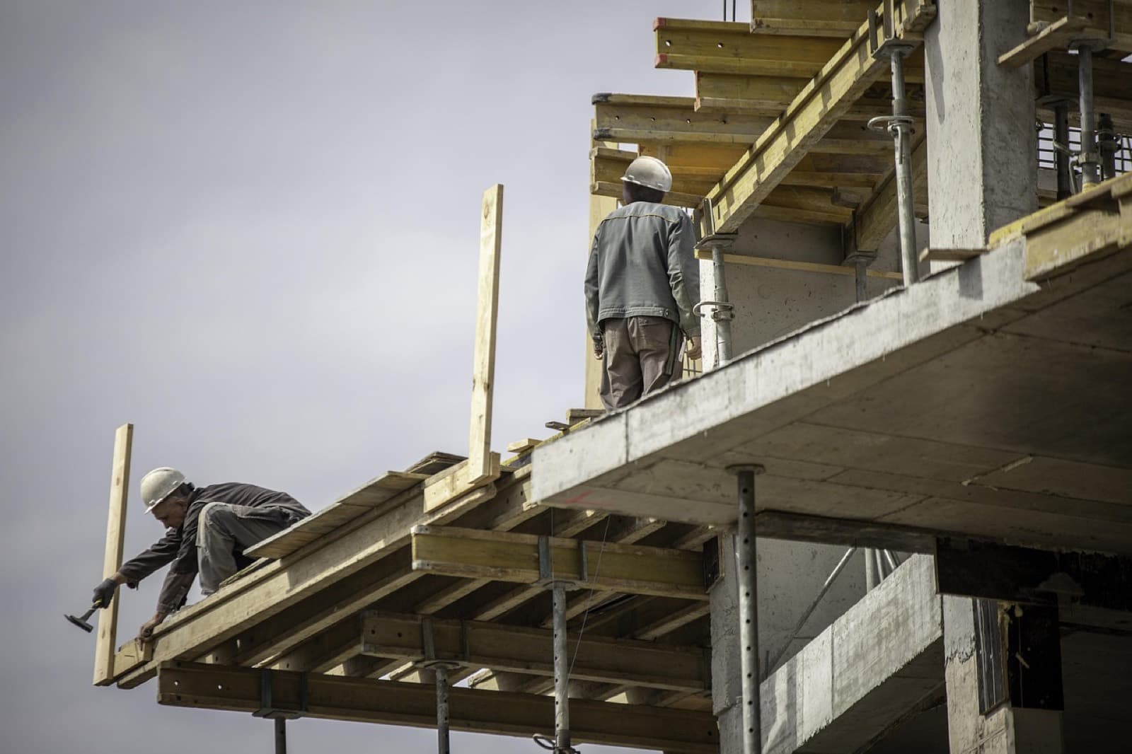 Due operai con il casco su un solaio in costruzione: uno fissa con il martello una tavola della cassaforma, l'altro sta in piedi poco più in là
