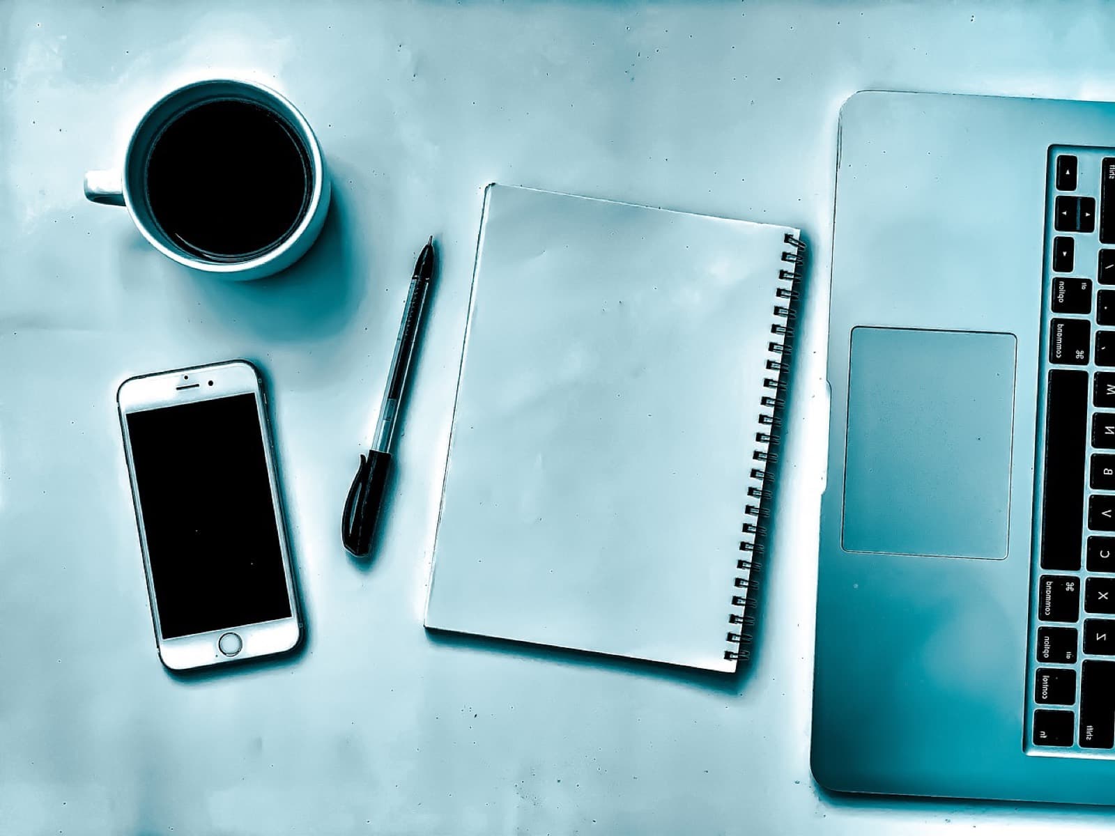 Una scrivania con un portatile aperto, un quaderno, una penna, il telefono e una tazza di caffè